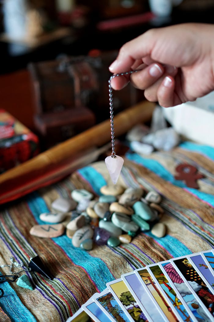 A hand holds a pendulum over a collection of tarot cards and stones, suggesting a mystical or spiritual setting.